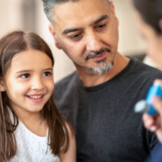 A parent listens as a clinician shows a young girl how to use an inhaler, illustrating supportive asthma education for families.