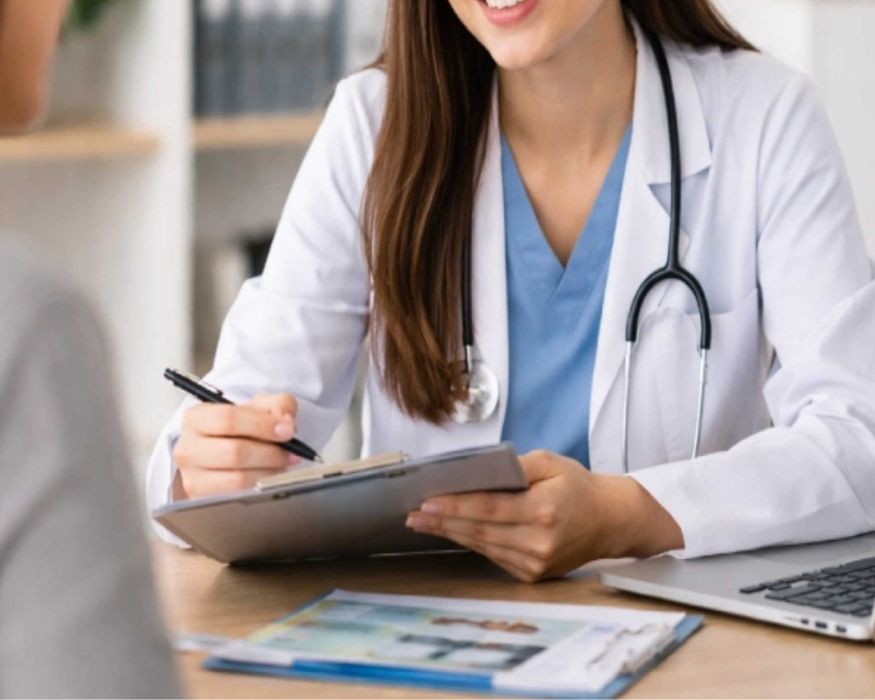A clinician smiles while taking notes on a clipboard, inviting patients to share their health concerns during the visit.