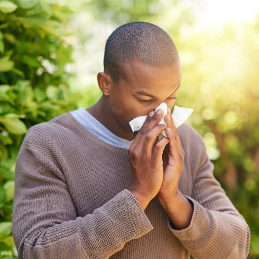 A man stands outdoors holding a tissue near his nose as he prepares to sneeze, suggesting seasonal allergy symptoms.