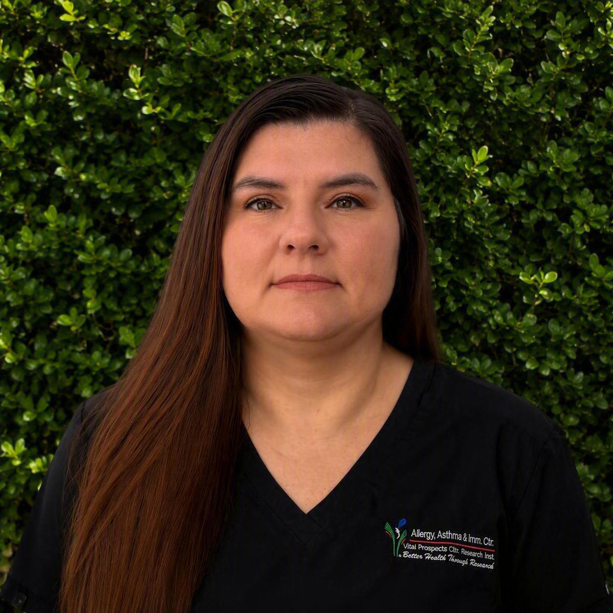 A member of the research team in black scrubs stands in front of green bushes, representing dedication for research and healing patients.