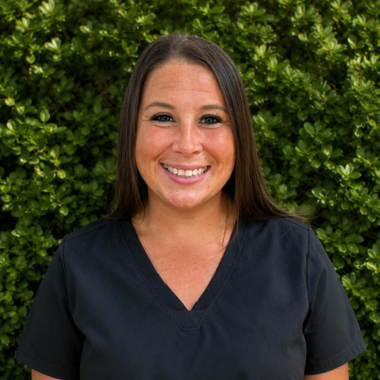 A smiling staff member in black scrubs stands outdoors before green foliage, presenting professional support in patient care.