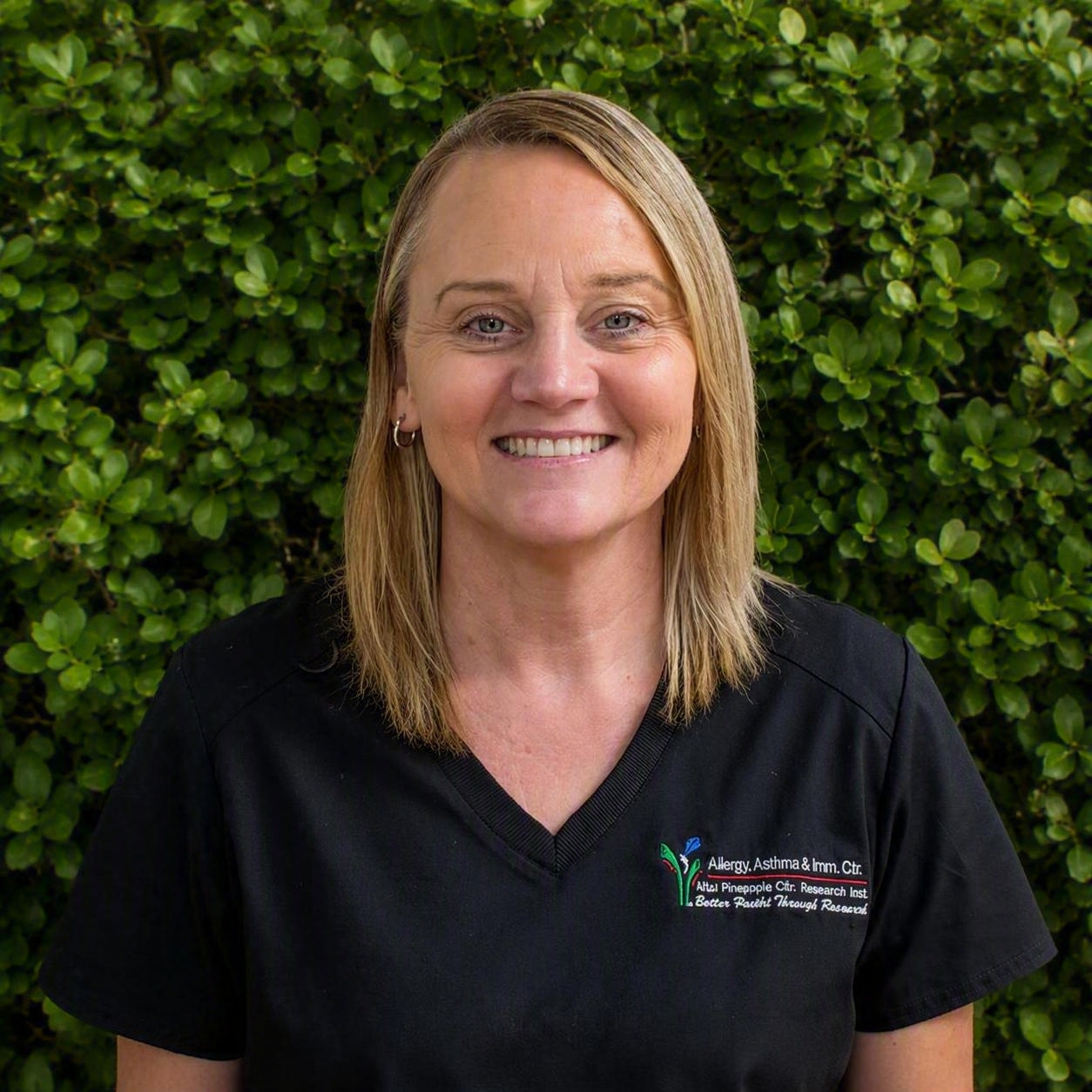 A clinical staff member in black scrubs stands in front of green shrubs, representing caring support for allergy and asthma patients.
