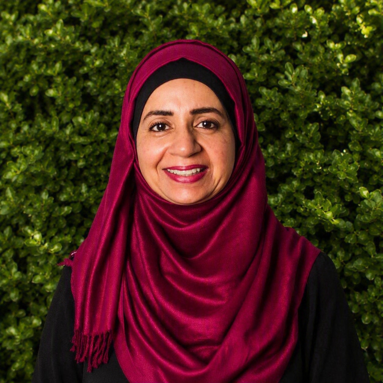 A team member wearing a deep red hijab stands outdoors against greenery, reflecting dedicated support in allergy and immunology services.