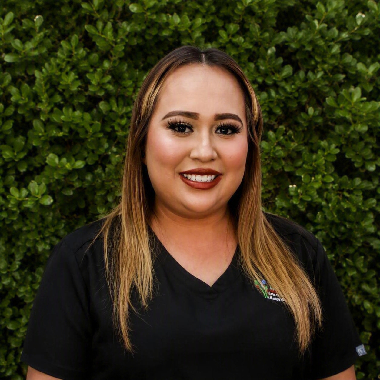 A clinical staff member in black scrubs stands in front of green shrubs, representing hands‑on support for allergy and asthma patients.