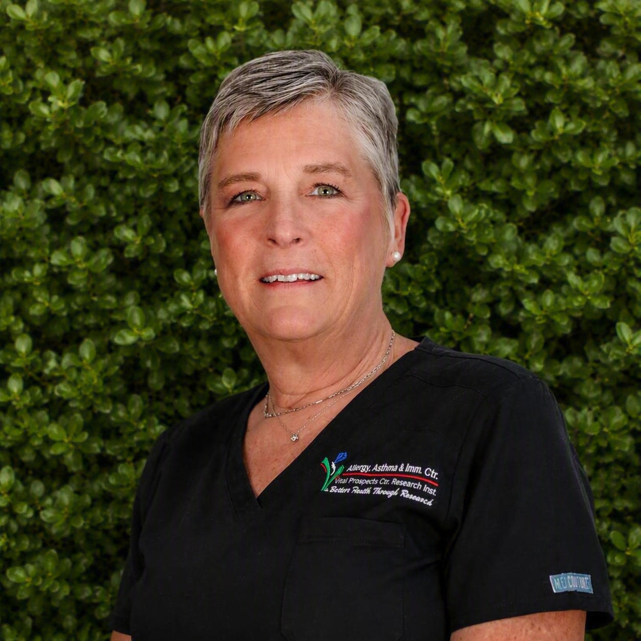 A medical staff member in black scrubs stands outdoors before green foliage, reflecting professional support in allergy and immunology care.